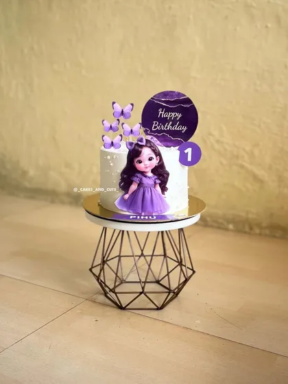 A simple and sweet first birthday cake for a baby girl, featuring a cute doll topper and purple butterflies.