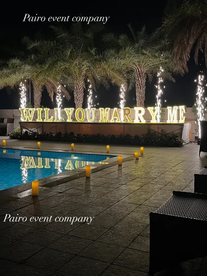 A dramatic poolside proposal at night. The setup includes large "WILL YOU MARRY ME" marquee letters reflecting in the water, a candle-lined walkway, and sparkling pyrotechnics for a truly memorable moment.