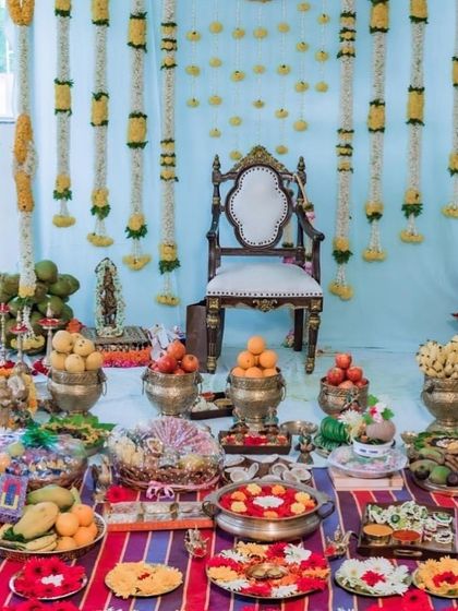 A very traditional and authentic baby shower setup, perfect for a home function. The decor uses brass vessels for the plate display and a simple yet elegant floral backdrop.