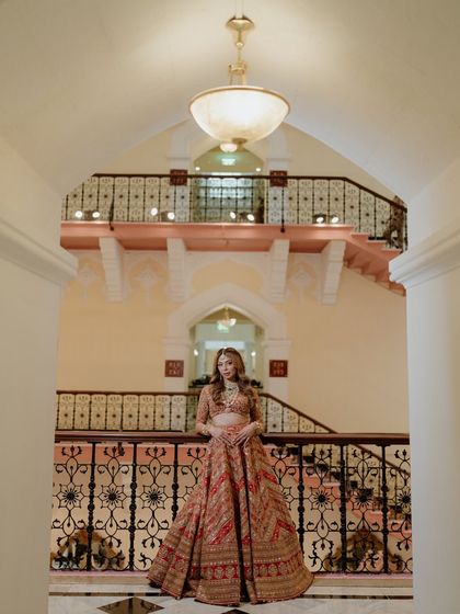 A full-length portrait of the bride standing on a balcony of the Taj Mahal Palace. The intricate ironwork and grand architecture frame her beautifully.