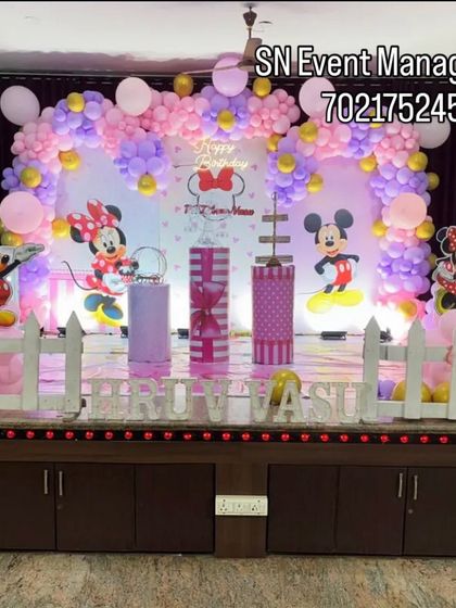 A cute and classic Minnie and Mickey Mouse stage decoration. The setup includes a picket fence, character cutouts, and a pink and purple balloon arch, perfect for a toddler's birthday.
