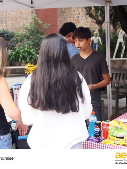 Students engage with their peers at a fundraiser table, explaining the cause they are supporting. This is an authentic way for them to practice advocacy and communication skills.