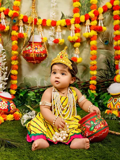 A little girl dressed as Radha sits amidst a colorful setup of marigold garlands and painted pots.