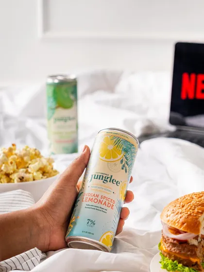 This lifestyle shot connects the product with relaxation and entertainment. A can of Junglee's Indian Spiced Lemonade is featured in a cozy "Netflix and chill" setup with a burger and popcorn.