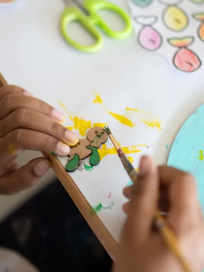 A close-up of tiny hands painting a small wooden cutout. In my classes, I focus on the process, encouraging kids to explore different brush strokes and color combinations. Every detail is a step in their creative journey.