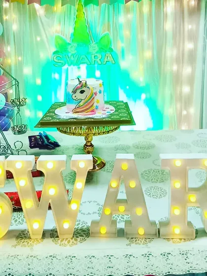 A magical unicorn-themed cake table setup for Swara's birthday. The display includes a custom cake, marquee letters, and a backdrop with fairy lights.