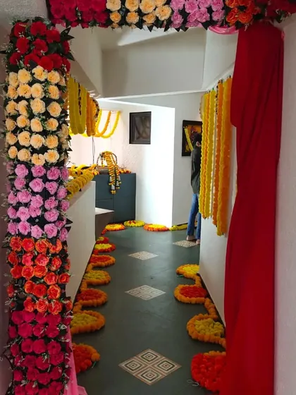 A vibrant entrance decoration for a home event like a haldi ceremony or welcoming the bride. The doorway is framed with colorful artificial flowers and a rich red drape, leading into a hallway decorated with marigolds.