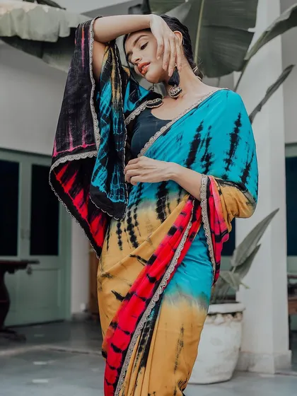 A model strikes a graceful pose in a vibrant tie-dye saree. The lush green background makes the blue, yellow, and red colors of the saree pop, creating a stunning image for a contemporary saree brand.
