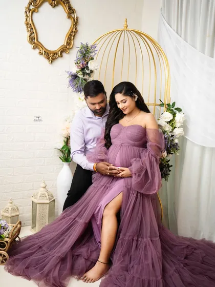 A full-length portrait showing the magnificent purple gown against the elegant studio setup. The pose highlights both the baby bump and the grace of the mother-to-be.
