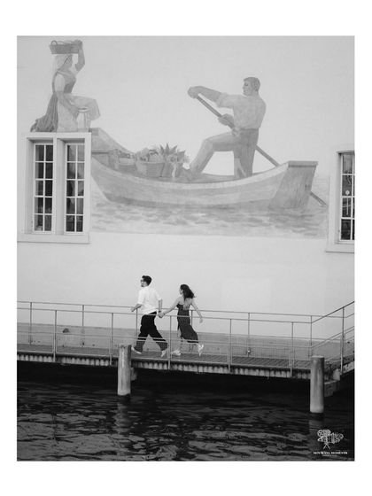 A black and white photo of a couple walking along a pier in Italy. A mural of a gondolier on the wall behind them adds a layer of artistic, old-world charm to the scene.