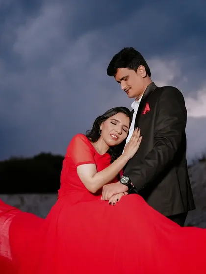 A tender moment against a dramatic, cloudy sky. The flowing red gown adds a vibrant splash of color to the moody landscape, creating a powerful and emotional image.