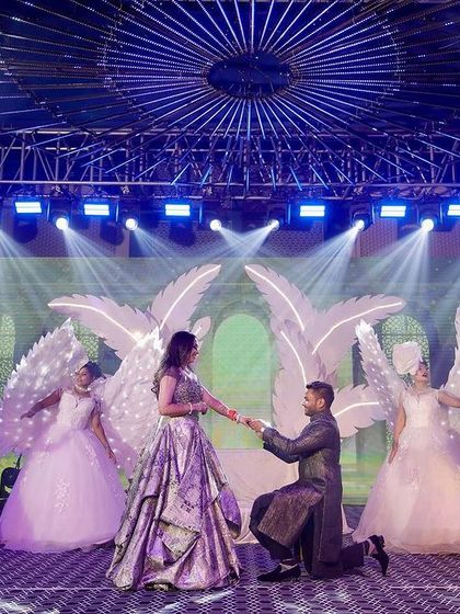 A theatrical Sangeet moment, where the couple is surrounded by performers in angel wings on a stage with dramatic lighting and a forest-themed LED backdrop.