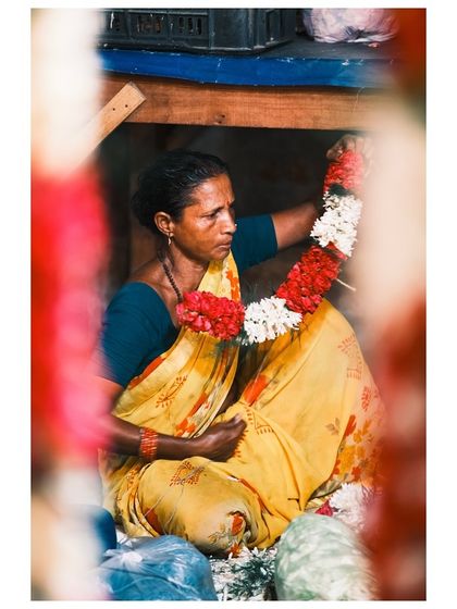 7:29am: Framed between colorful garlands, a woman in a yellow sari weaves flowers at the market. This shot uses a natural frame to draw the viewer's eye to her work.