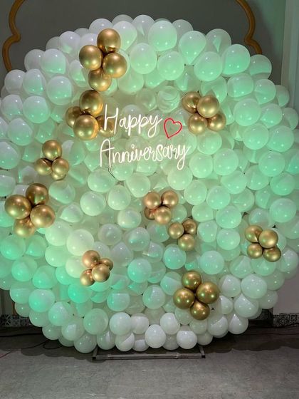 This stunning anniversary backdrop features a full wall of white balloons with elegant gold balloon accents. The "Happy Anniversary" neon sign in the middle glows beautifully, creating a sophisticated and celebratory focal point.