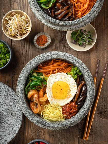 A Japanese hot pot, or Bibimbap, with shrimp and a fried egg. This top-down view for YouMee captures the colorful arrangement of ingredients in the stone bowl.