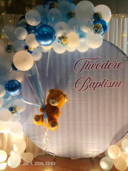 A charming teddy bear themed backdrop for a Baptism ceremony. This setup features a custom printed circular backdrop with the child's name, framed by a blue and white balloon arch, with a cute teddy bear appearing to float with balloons.