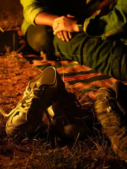 The end of a perfect day. Their shoes rest on the ground by a picnic blanket, with a bonfire's glow in the background. This detail shot tells a story of rest and contentment after an adventure.