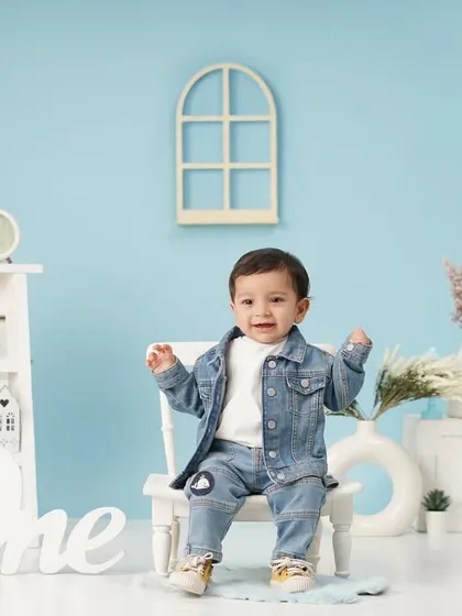 A happy baby boy in a denim-on-denim outfit smiles for his first birthday photo, surrounded by a light blue and white set.
