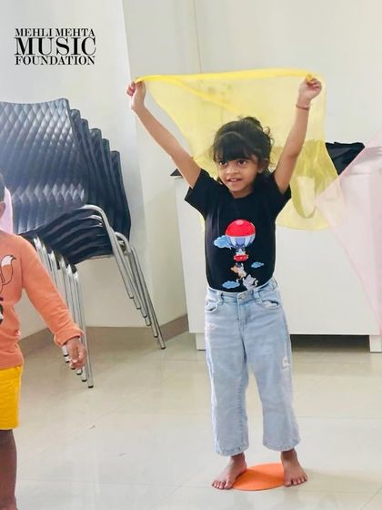 A young girl joyfully waves a yellow scarf, participating in a movement activity designed to teach musical expression. Our classes are full of color, energy, and smiles.