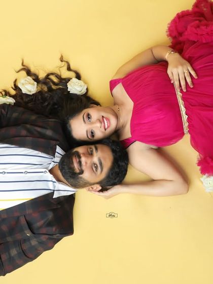 A creative and romantic top-down shot of an expecting couple. The vibrant pink gown and scattered white roses on the yellow background make for a unique and colorful portrait.