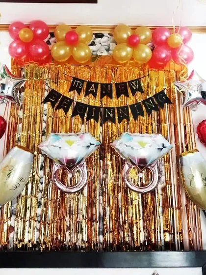 An engagement party or bridal shower backdrop. This fun setup features a gold shimmer curtain, 'It's a New Beginning' banner, and large foil balloons shaped like diamond rings and champagne glasses.