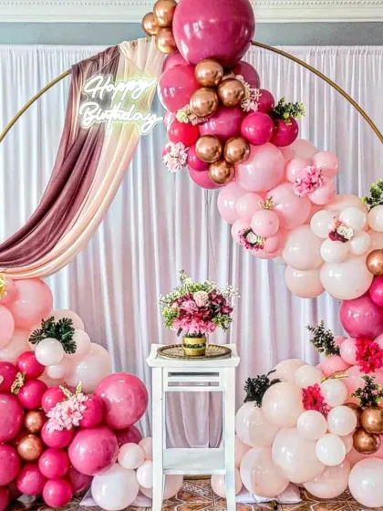 This stunning birthday backdrop features a gold ring frame draped with fabric and a lush balloon garland in shades of pink, rose gold, and white. It's a very popular choice for elegant home parties.