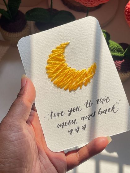 A hand-held shot of the "I love you to the moon and back" embroidered card. The yellow thread really pops against the white paper.