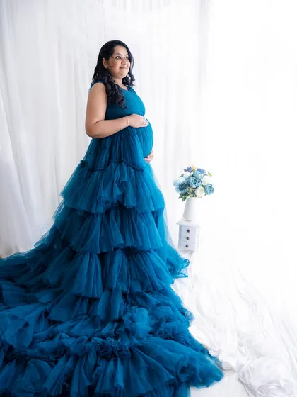 A stunning full-length portrait of a mom-to-be in a dramatic, tiered blue gown against a bright, clean background.