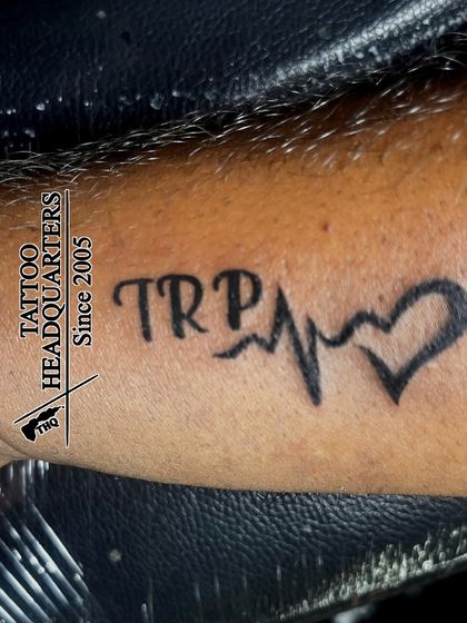 A tattoo combining the initials "TRP" with a heartbeat line (EKG) that forms a heart at the end.