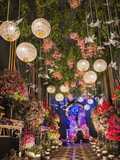 This is an enchanted forest themed entrance passage. The design uses hanging greenery, pink flowers, paper bird ornaments, and woven lanterns to create a whimsical and unique path for guests.