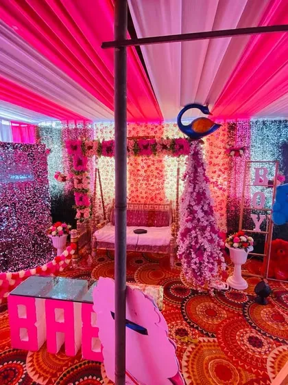 A wide view of a baby shower pandal, showing the stage with a moon prop, sequin wall, and decorated swing under a pink and white ceiling.
