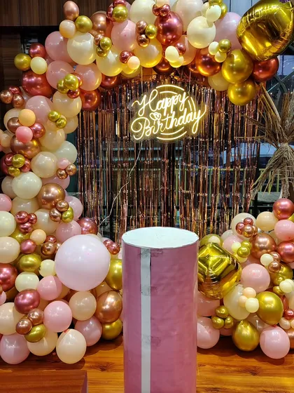 A glamorous birthday backdrop with a rose gold, pink, and gold balloon arch against a metallic fringe curtain.