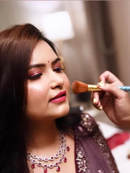 This behind-the-scenes shot captures the final touches of a dewy makeup look for a Roka ceremony. The focus is on fresh, glowing skin, complemented by soft pink eyeshadow and a beautiful diamond and ruby necklace.