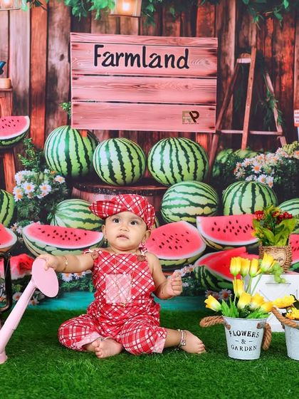 A baby girl sits in a charming "Farmland" setup, surrounded by watermelons and flowers. The rustic props and cheerful red outfit make for a unique and adorable portrait.