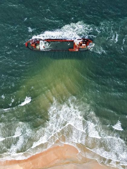 A top-down aerial photograph of the abandoned ship at Suratkal Beach, Mangalore, showing the contrast between the rusty vessel and the green-blue water.