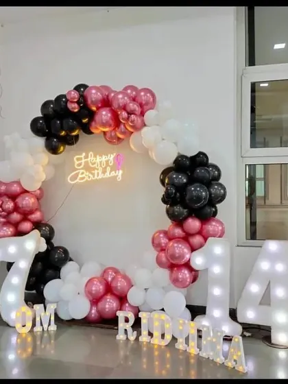 This decoration for a 14th birthday uses a trendy color combination of pink, black, and white. The setup includes a circular balloon arrangement, light-up numbers, and the birthday girl's name in light-up letters for a personalized touch.