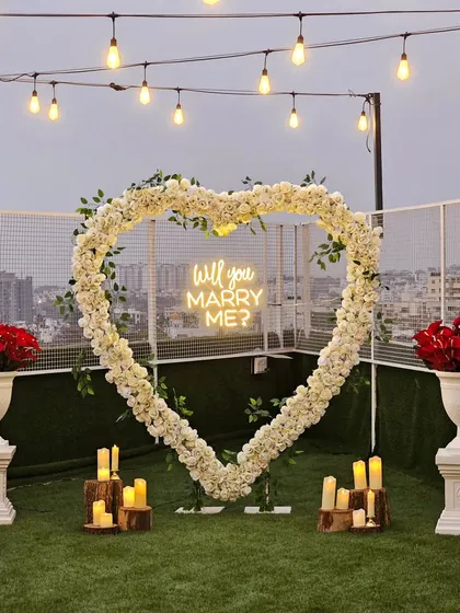 A chic and modern rooftop proposal setup with a stunning city view. The focal point is a large heart-shaped arch of white flowers and greenery, accented with a "Will you Marry Me?" neon sign and surrounded by candles on rustic wood stumps.