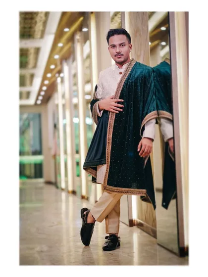 A stylish portrait of the groom in his royal attire. He leans against a mirrored wall, looking confident and dapper in his cream kurta and green velvet shawl.