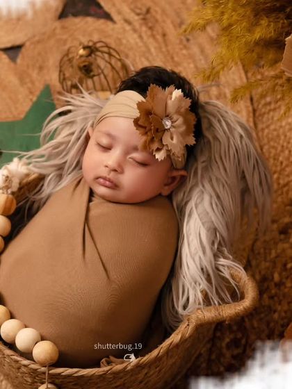 A close-up, top-down view of a newborn wrapped in a brown swaddle, nestled in a basket with a large, beaded garland. The soft focus on the edges draws attention to the baby's peaceful face.