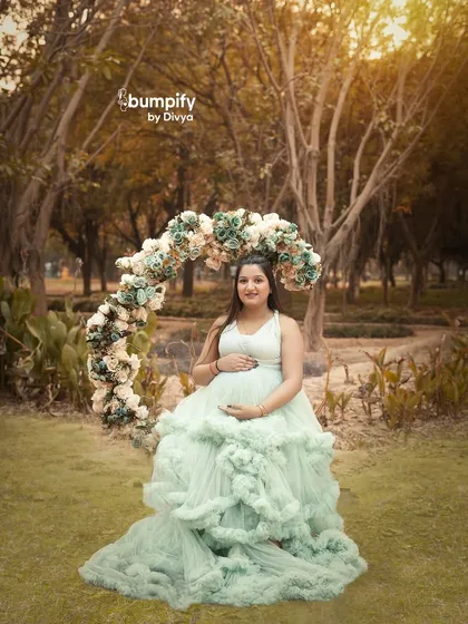 A dreamy outdoor portrait with a beautiful floral arch. The mom-to-be is seated, surrounded by flowers, wearing a lovely light green gown that complements the natural setting.
