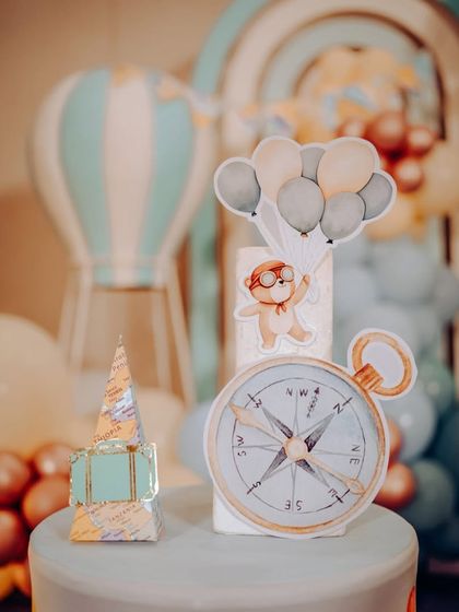 A close-up of a travel-themed cake topper, featuring a compass, a suitcase, and a bear floating with balloons.