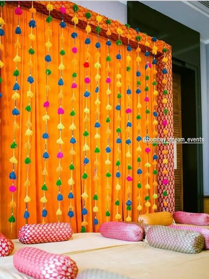A simple yet vibrant backdrop for a home Mehndi or Dholki, using colorful pom-pom strings against a bright orange drape.
