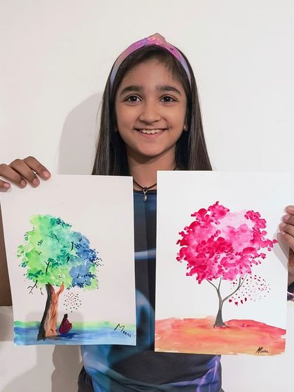 A student displays two of her beautiful tree paintings, one with a meditating figure and another with vibrant pink blossoms.