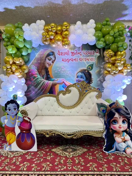 A grand baby shower stage with a large Radha Krishna backdrop. The setup is framed with green and white balloons and includes a sofa for seating.