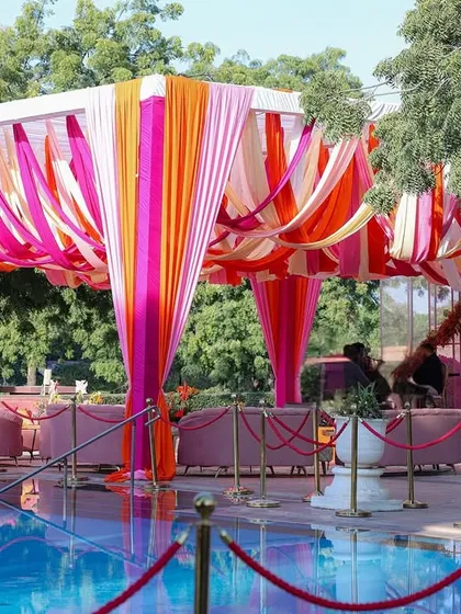 A vibrant poolside seating area with lounges set under a canopy of pink and orange hanging drapes, perfect for a high-tea event.