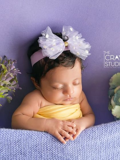 A sleeping baby girl wearing a large, delicate bow, nestled between soft floral props. The combination of yellow and purple creates a sweet, gentle color palette.