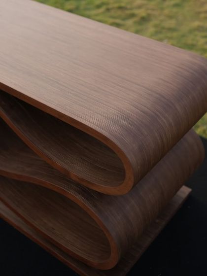 A close-up view of the SWISH bench, highlighting the smooth curves and the rich texture of the wood veneer. The layers of plywood are visible at the edges, revealing the honesty of its construction.