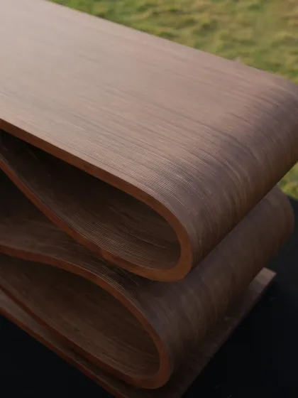 A close-up view of the SWISH bench, highlighting the smooth curves and the rich texture of the wood veneer. The layers of plywood are visible at the edges, revealing the honesty of its construction.