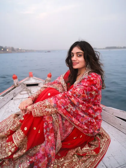 A model smiles warmly while sitting in a boat on the Ganga, dressed in a vibrant red traditional outfit. This image captures the pure joy and serene beauty of a classic Banaras experience.