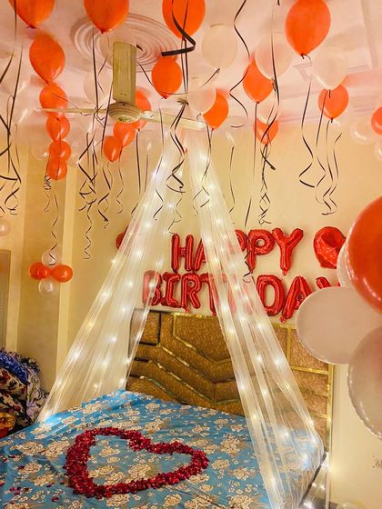 A warm and inviting birthday bedroom setup featuring a fairy light canopy, a heart of rose petals, and a ceiling full of orange and white balloons.